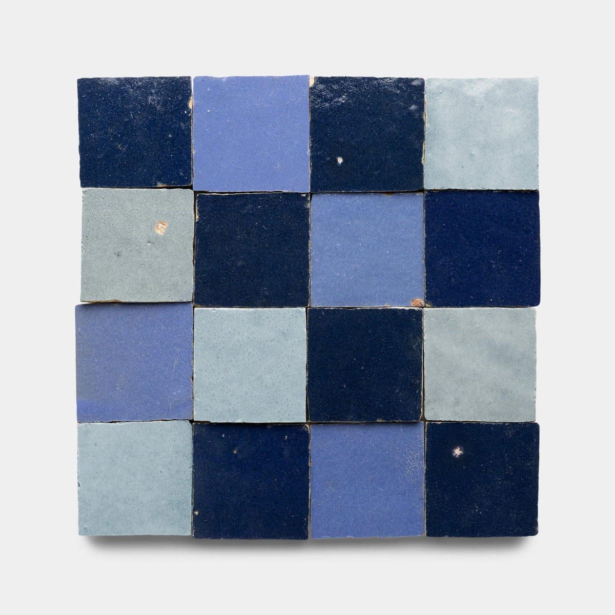A square grid of Zia Tile’s Night Blue + Skylight + Portuguese Blue Checkerboard sample displays sixteen handmade tiles with unique imperfections, arranged in a checkerboard pattern on a white background.