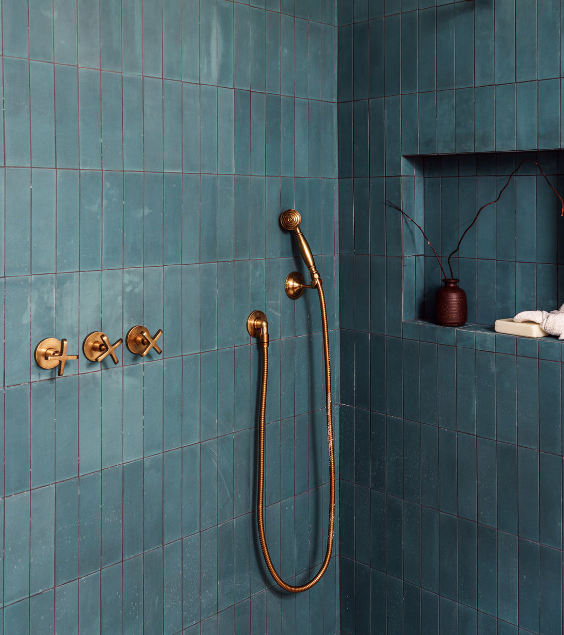 A modern shower features vendor-unknown's Ocean 2x8 subway tile, gold fixtures, a handheld showerhead, and a recessed shelf displaying a brown vase with twigs and neatly stacked white towels.