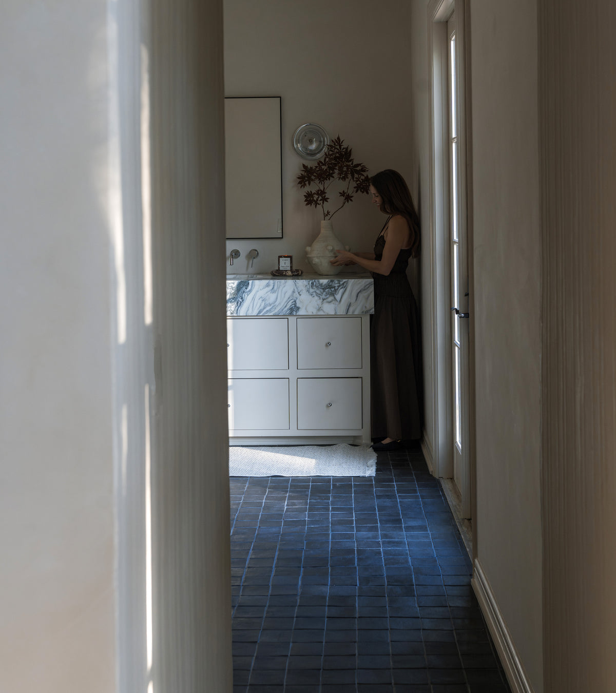 A woman arranges flowers at a marble sink in a softly-lit hallway with light walls and handmade 4x4 Square + Oscura tiles by Zia Tile.