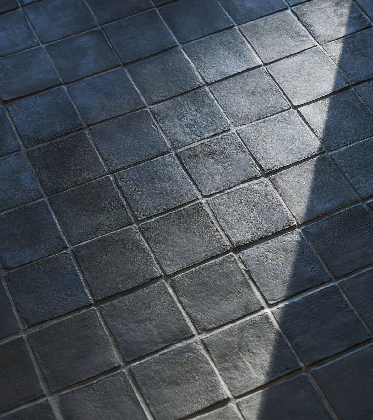 Close-up of a dark tiled floor featuring Zia Tile’s 4x4 Square + Oscura handmade Cotto tiles. Light and shadow highlight the textured, uneven ceramic surface arranged in a grid pattern.