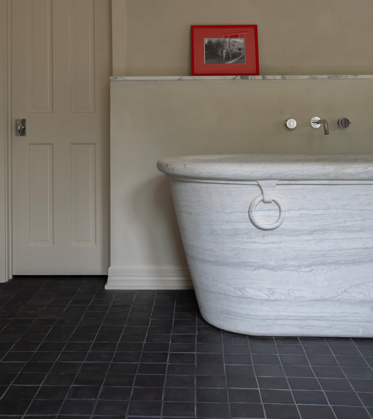A modern bathroom with a large marble bathtub, beige walls, a light door, and a small shelf with a red-framed photo above the tub features Zia Tile's 4x4 Square + Oscura handmade black Cotto tiles on the floor.