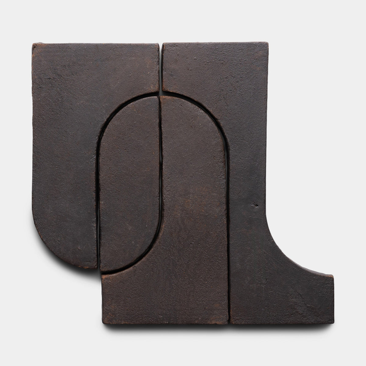 The Durango + Oscura, Sample by Zia Tile is a dark, matte-finished abstract ceramic piece with two rectangular forms featuring curved cutouts, creating a minimalist geometric design against a light background.
