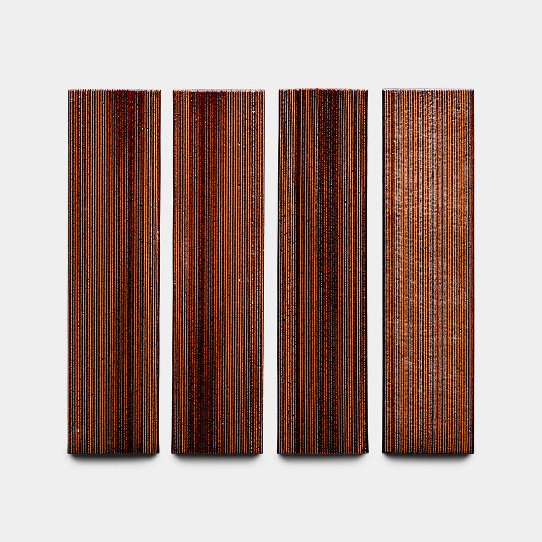 Four rectangular Oxblood + Etched panels by Zia Tile, each with a textured dark brown surface and fine vertical lines, are evenly spaced against a plain light gray background.
