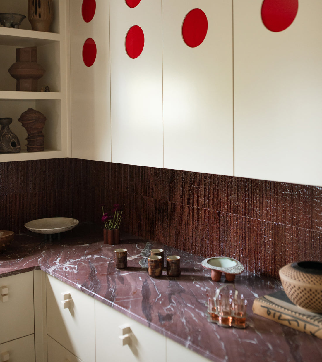 A kitchen counter with a dark red marble surface and a textured Oxblood + Furrowed tile backsplash by Zia Tile, featuring cream cabinets with large red circular accents.