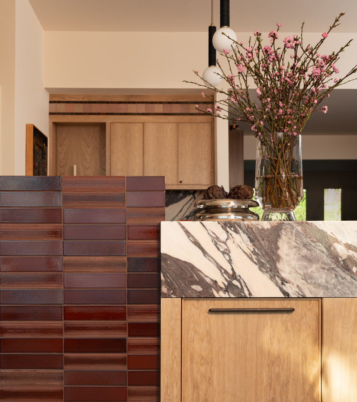 A wall section of Oxblood + Etched rectangular tiles next to a kitchen island with a marble countertop and wooden cabinets.