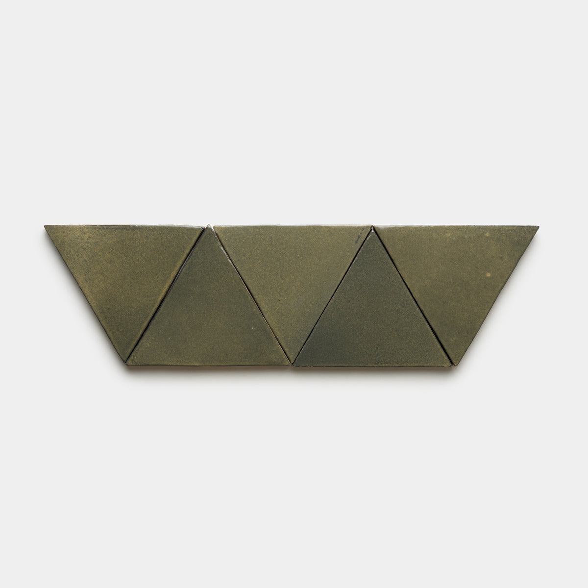 Four Oyamel Triangle tiles by Zia Tile, in a greenish-gray hue, are arranged in a row with two triangles at the center and two trapezoids at each end, set against a plain white background.