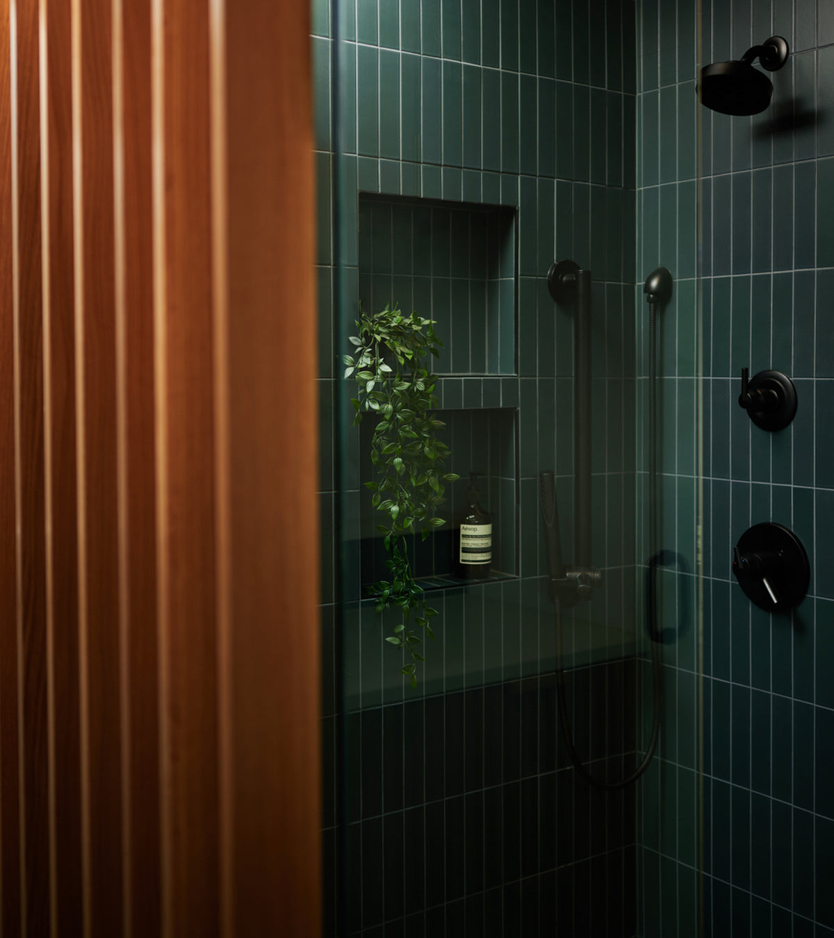 A modern shower showcases Zia Tile’s Pacific Blue 2x8 ceramic tiles set vertically, with black fixtures and a built-in shelf holding a plant and bottle, partially framed by vertical wooden slats on the left.