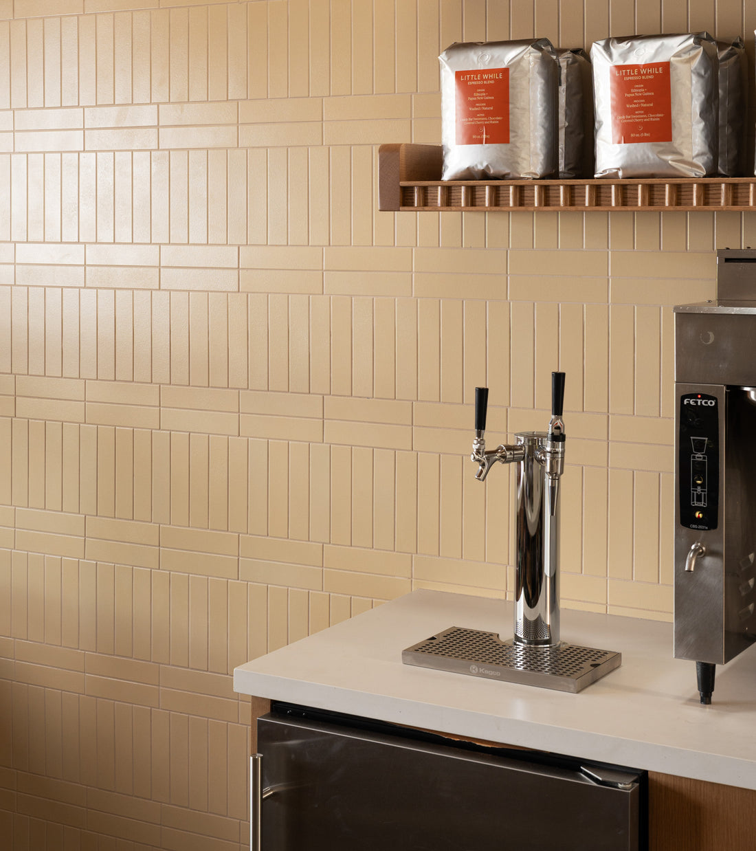 A modern coffee station with a white countertop, chrome coffee tap, stainless steel machine, and two large coffee bags on a wooden shelf against a Pale Ochre 2x8 Zia Tile wall.