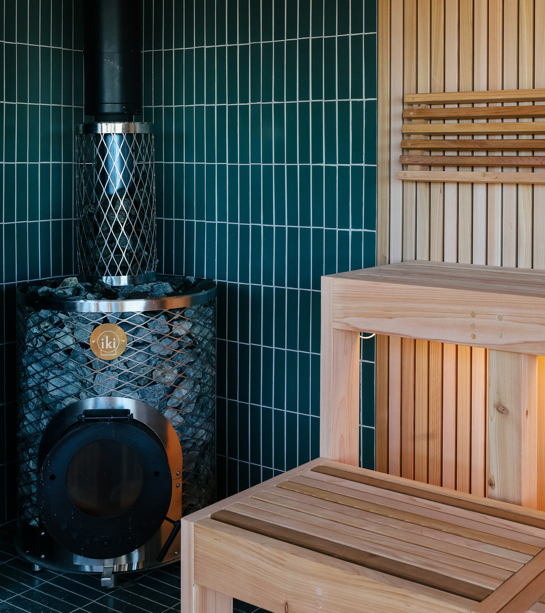 A modern sauna interior with a wooden bench, slatted wood wall, and black metal sauna heater with glass door and rocks, set against dark green Patina 2x8 tiles by Zia Tile.
