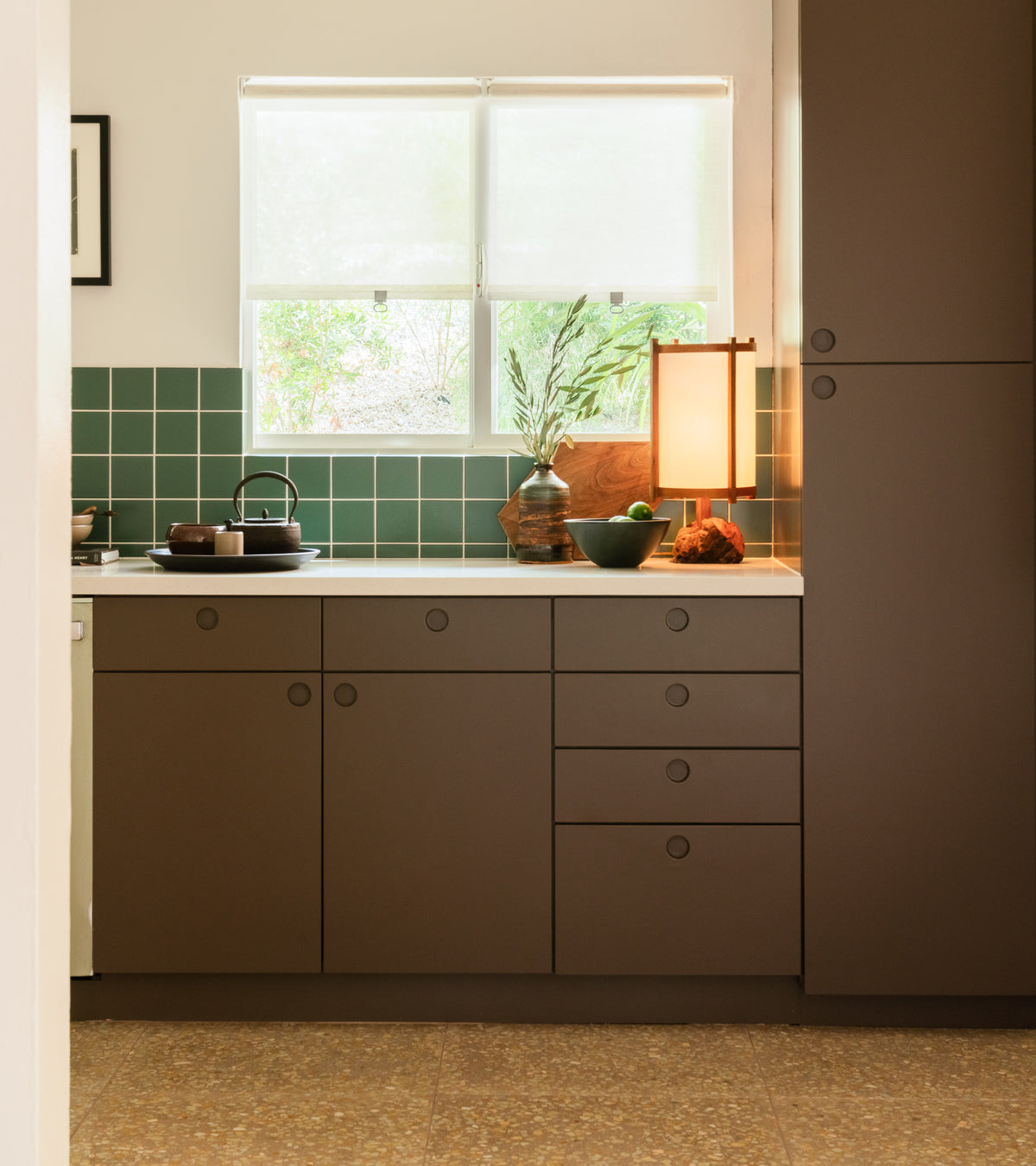 A modern kitchen features brown cabinets and a green Zia Tile Patina 4x4 handmade tile backsplash, a countertop with fruit, teapot, and decor, all under a window with white blinds letting in natural light.