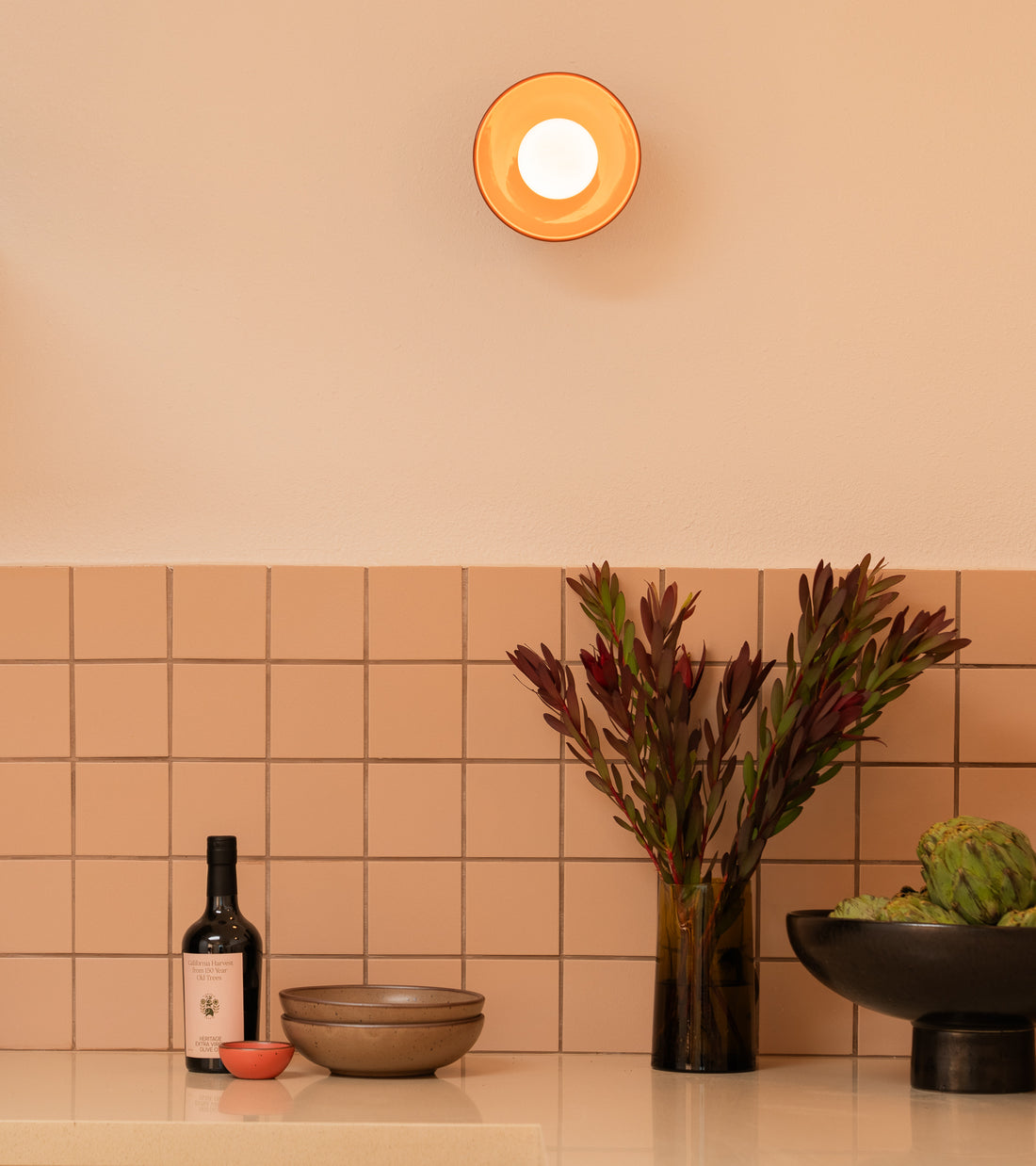 A modern kitchen counter with Zia Tile’s Peach Blossom 4x4 handmade tile, a bottle, bowls, a small red dish, a vase with dark green and red foliage, artichokes in a black bowl, and a round wall light above.