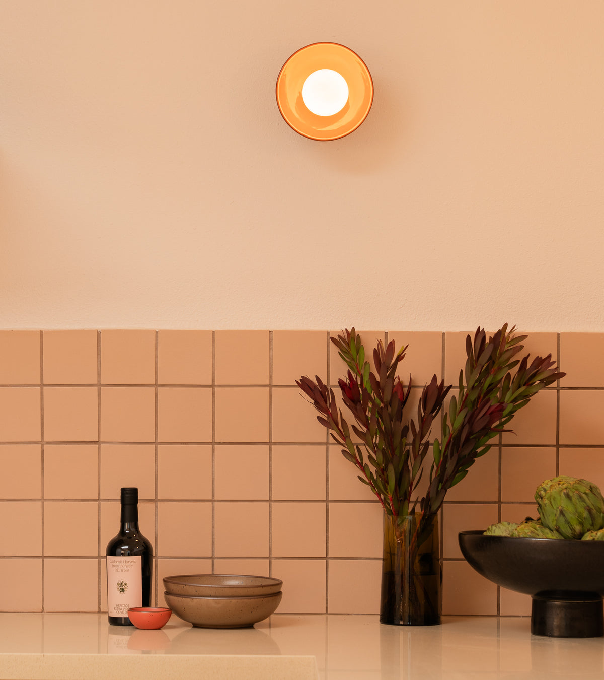 A modern kitchen counter with Zia Tile’s Peach Blossom 4x4 handmade tile, a bottle, bowls, a small red dish, a vase with dark green and red foliage, artichokes in a black bowl, and a round wall light above.