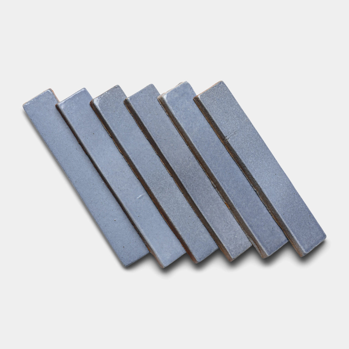 Six Pedregal Mini Bar tiles by Zia Tile, in a blue-gray hue, are arranged in a staggered, overlapping pattern on a white background.