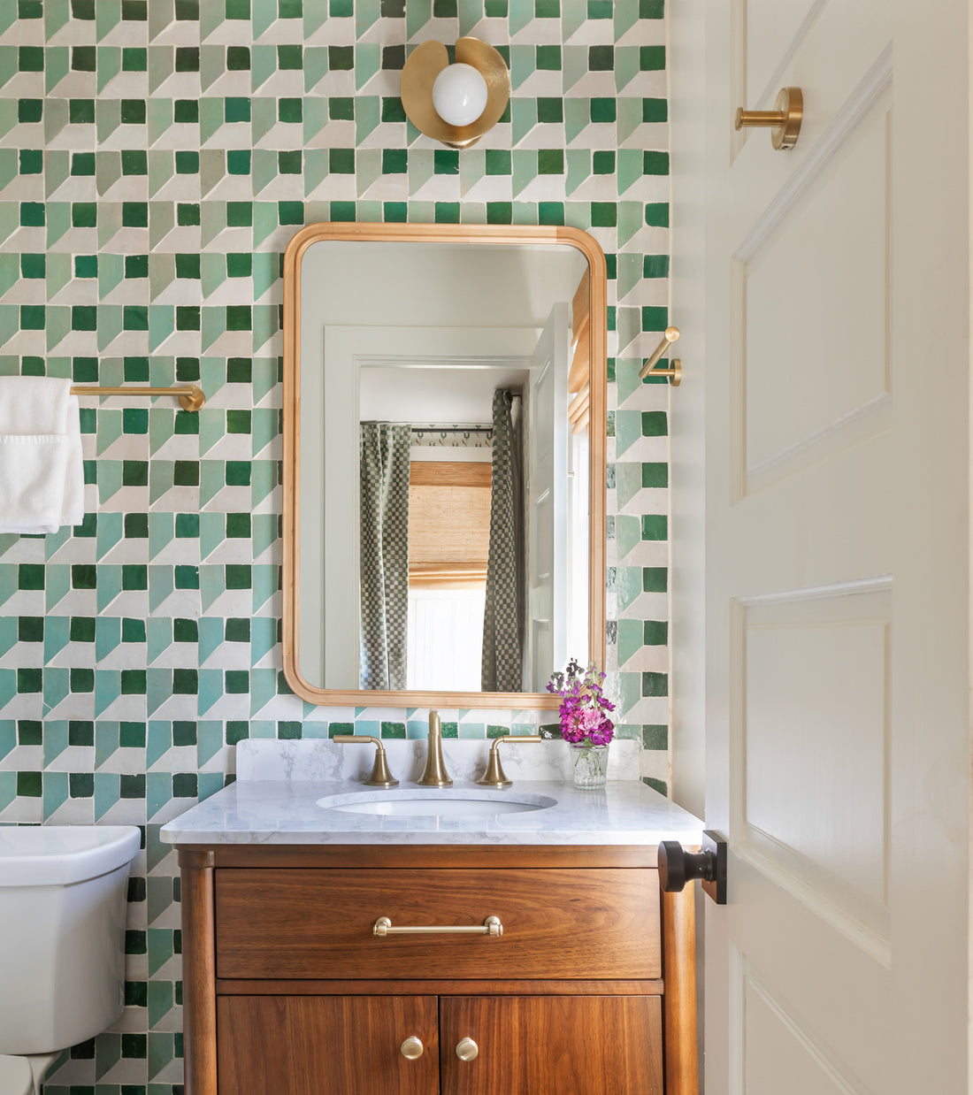 A bathroom featuring a Perpetual Check 2 green and white Moroccan tile wall by Zia Tile, wooden vanity with marble top, gold faucet, rectangular mirror, wall sconce, towel bar, and a small vase of purple flowers.