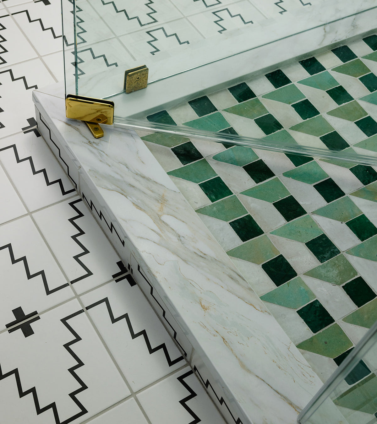 Close-up of a shower entryway with a glass door, green and white Moroccan tile inside, white marble trim, and Zia Tile’s Perpetual Check 2 black-and-white floor tiles with zigzag patterns outside the shower.