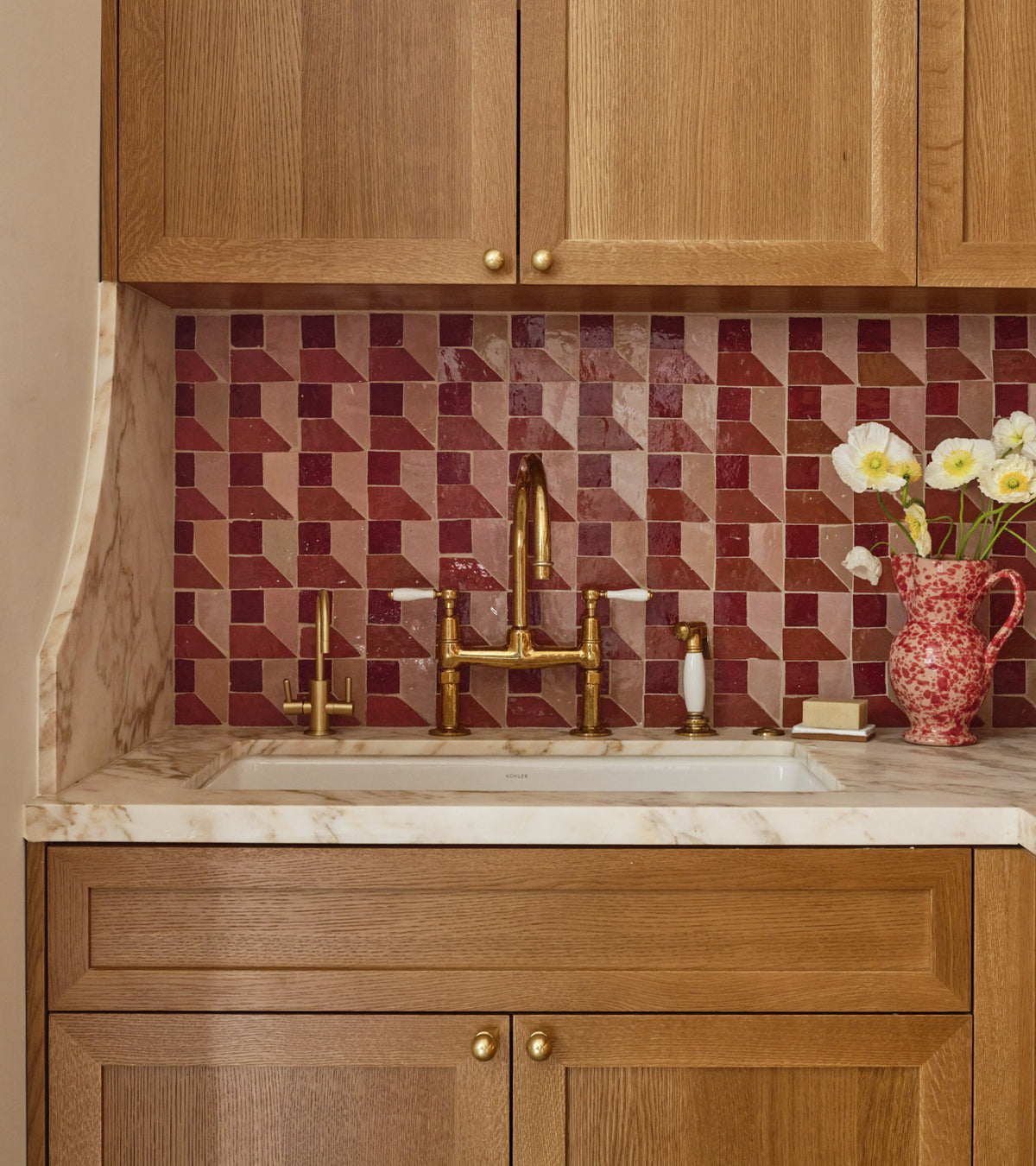 A kitchen sink area with wood cabinets, a marble countertop, and a backsplash of Zia Tile's Perpetual Check 3 red and pink Moroccan zellige tiles. Gold fixtures and a red patterned vase with white flowers complete the charming look.