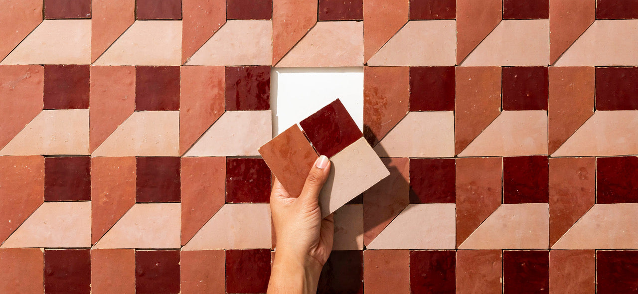 A hand holds two geometric tiles against a wall covered in similar tiles with a 3D cube pattern in shades of red, pink, and beige. One tile has been removed, revealing a white space underneath.