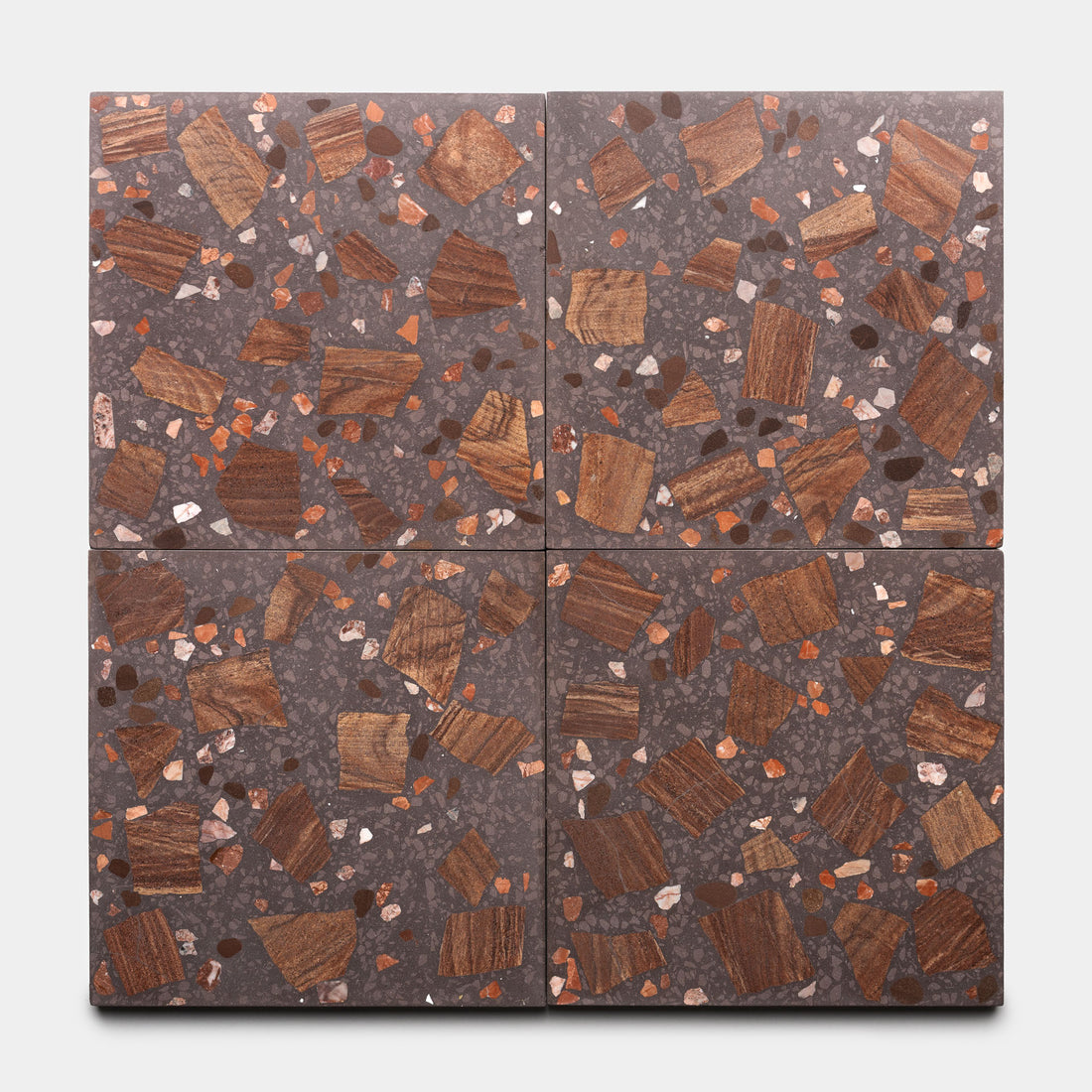 The Petrichor 12x12 tiles by Zia Tile feature a dark grey terrazzo base with irregular brown, rust, and beige stone and wood chips. Four tiles create a geometric pattern when arranged together.