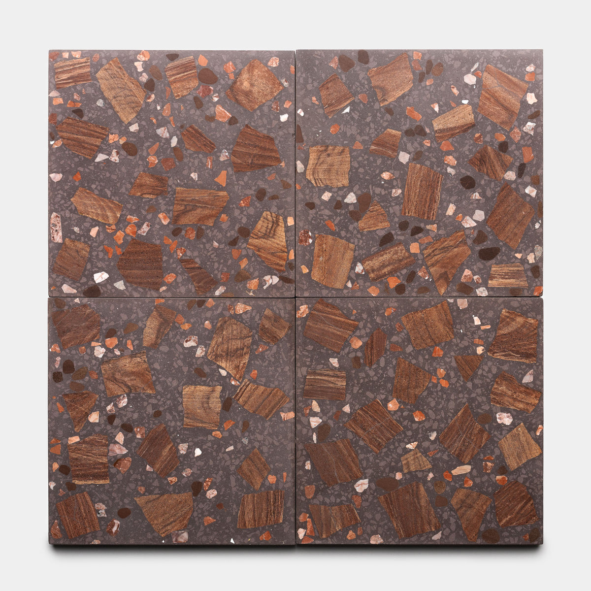 The Petrichor 12x12 tiles by Zia Tile feature a dark grey terrazzo base with irregular brown, rust, and beige stone and wood chips. Four tiles create a geometric pattern when arranged together.