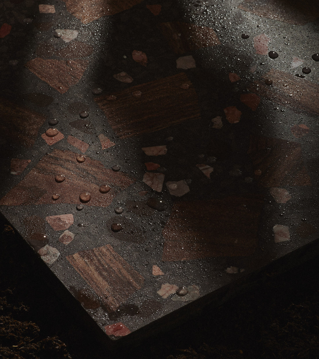 A close-up of Zia Tile’s Petrichor 12x12 terrazzo shows reddish and pinkish fragments, water droplets, and partial shadows on the surface. The edge is visible with soil below.