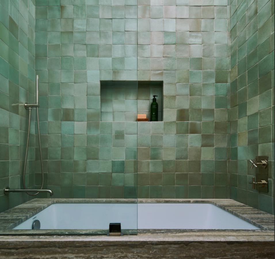 A modern bathroom features Zia Tile's Peyote 4x4 Square green handmade tiles, a built-in shelf with soap and a bottle, a marble-edged tub, and wall-mounted shower controls.