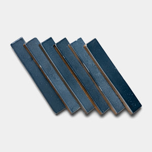Six rectangular Piedra Mini Bar tiles by Zia Tile, in dark blue ceramic, are arranged diagonally with slight overlaps on a light gray background.