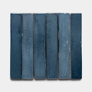 Six glossy navy blue Piedra Mini Bar tiles by Zia Tile are arranged vertically on a light gray background. Each rectangular ceramic tile shows subtle color and texture variations.