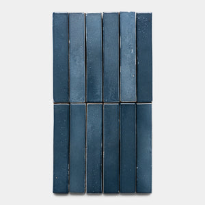 Twelve glossy, rectangular Piedra Mini Bar tiles by Zia Tile in dark blue, with subtle variations and speckles, are arranged vertically in three columns and four rows on a light gray background.