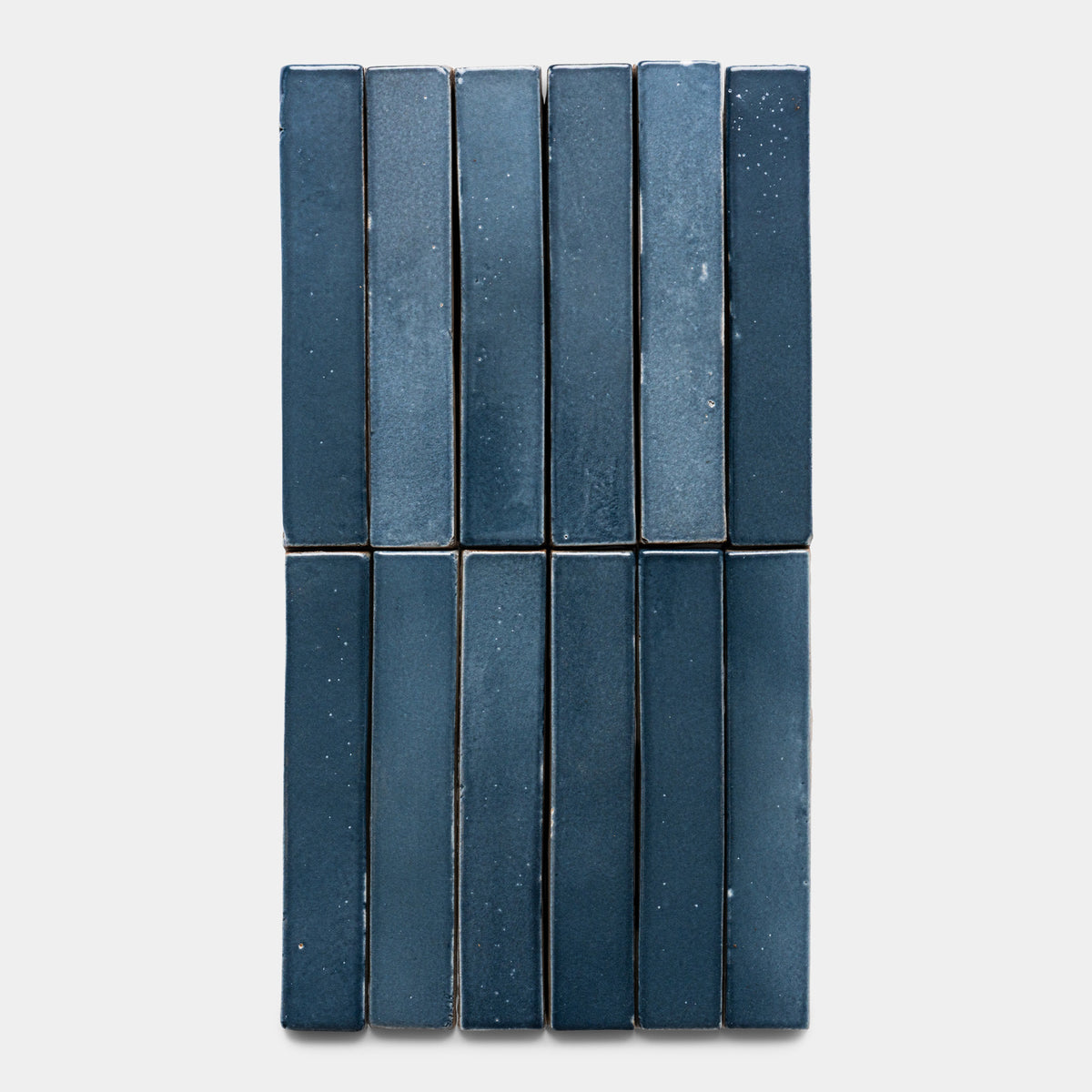 Twelve glossy, rectangular Piedra Mini Bar tiles by Zia Tile in dark blue, with subtle variations and speckles, are arranged vertically in three columns and four rows on a light gray background.