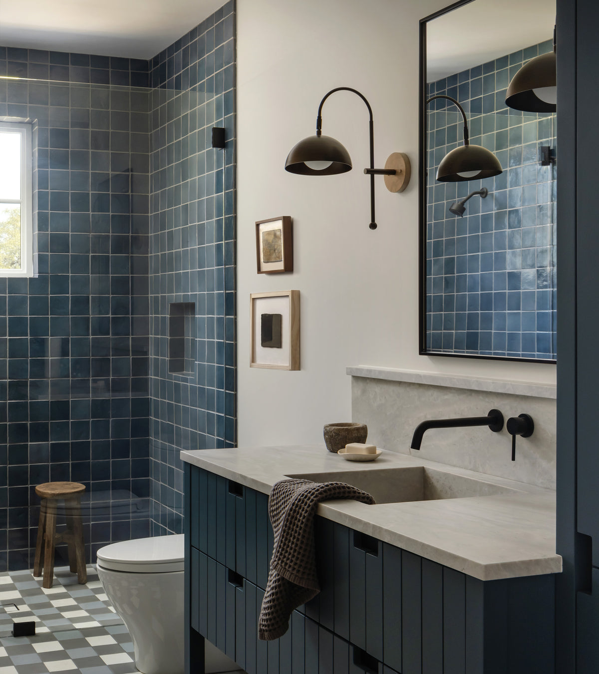 Modern bathroom featuring Zia Tile’s Piedra 4x4 Square blue handmade ceramic tiles in the shower, checkered floor, floating double vanity, black fixtures, large mirror, wall sconce, framed art, wooden stool, and natural light.