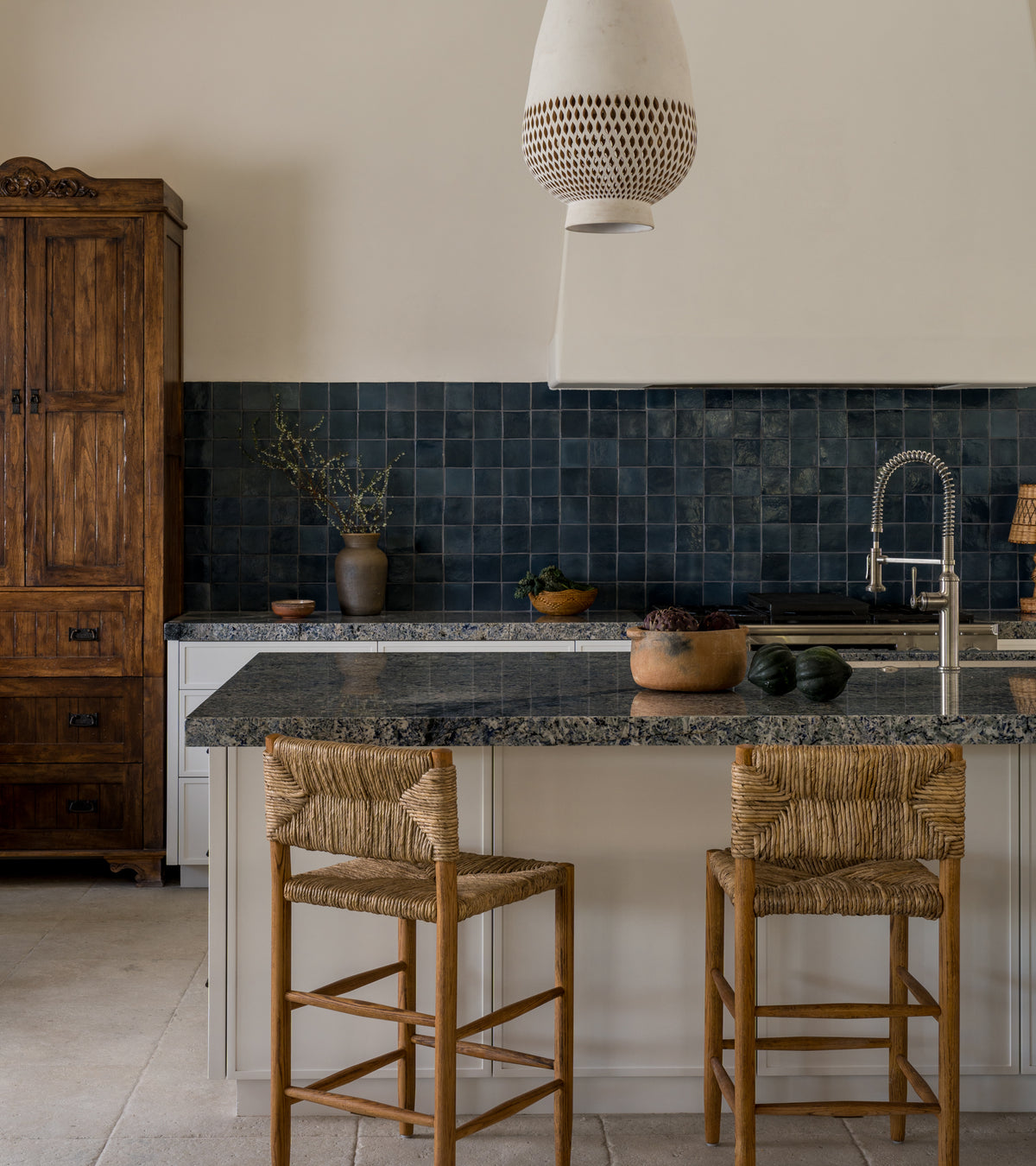 A modern kitchen features a Zia Tile Piedra 4x4 Square dark blue tile backsplash, a granite island with sink, two woven bar stools, wooden cabinets, ceramic-inspired pottery, and a white pendant light.