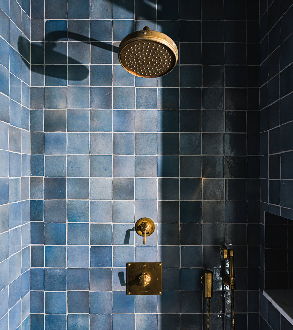 A shower featuring Zia Tile’s glossy blue Piedra 4x4 Square tiles and handmade Cotto accents, with a brass rain showerhead, matching controls, and a built-in niche for the handheld shower wand.