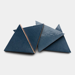 Four glossy, dark blue Piedra Triangle ceramic tiles by Zia Tile are arranged overlapping each other on a white background.