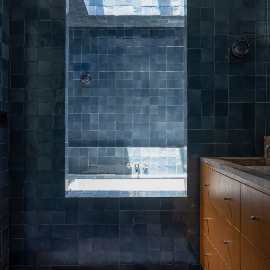 A modern bathroom showcases Zia Tile’s Piedra 4x4 Square tiles on the walls and floor, a built-in tub under a skylight, and a wooden vanity with drawers on the right. Natural light brightens the space.