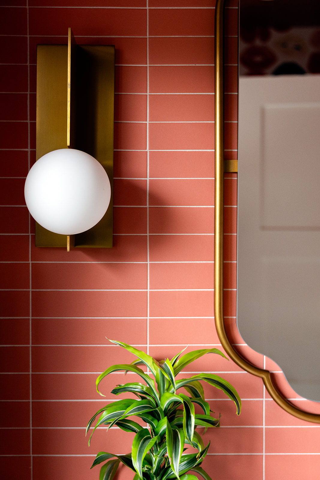 A round white wall sconce with a brass mount is attached to a wall of Zia Tile's Pink Dahlia 2x8 matte ceramic tiles. A gold-framed mirror and a green potted plant are visible in the foreground.
