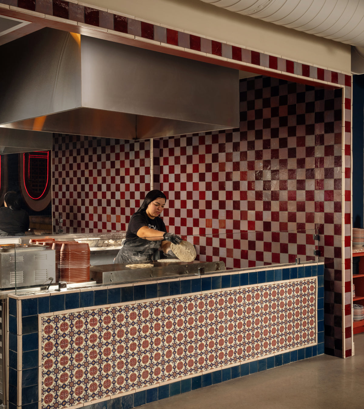 A worker in a black shirt and gloves prepares flatbread on a metal cooktop in front of a red and white checkered Plum 4x4 tile backsplash.