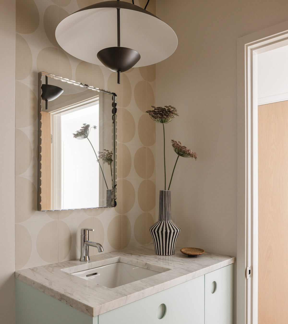 A modern bathroom features a rectangular marble sink, a silver-framed mirror, a tall vase with flowers, large-beige-circle wallpaper, a Pomelo Bone 8x8 tile floor by vendor-unknown, and a black-and-white ceiling light. The door is slightly open.