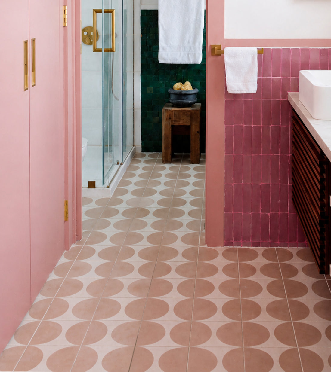 A modern bathroom featuring Pomelo Jaipur Pink 8x8 tile by vendor-unknown, geometric flooring, gold hardware, a glass shower, and a wooden stool with towels and soap bowl.