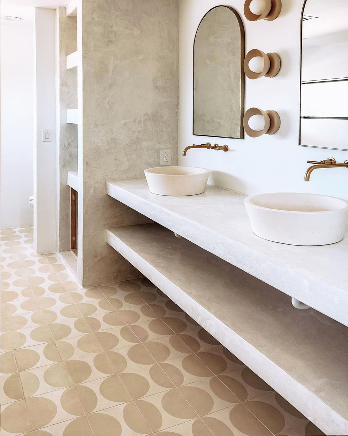 Modern bathroom with geometric Zia Tile Pomelo Bone 4x4 floor tiles, a double white concrete vanity with round vessel sinks, arched mirrors, minimalist gold faucets, and neutral walls create a clean, contemporary style.