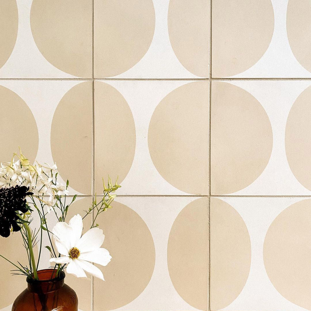 A brown glass vase with white and dark flowers sits in front of a wall tiled with Zia Tile's Pomelo Bone 4x4, Sample, featuring a beige and white geometric pattern of overlapping circles.