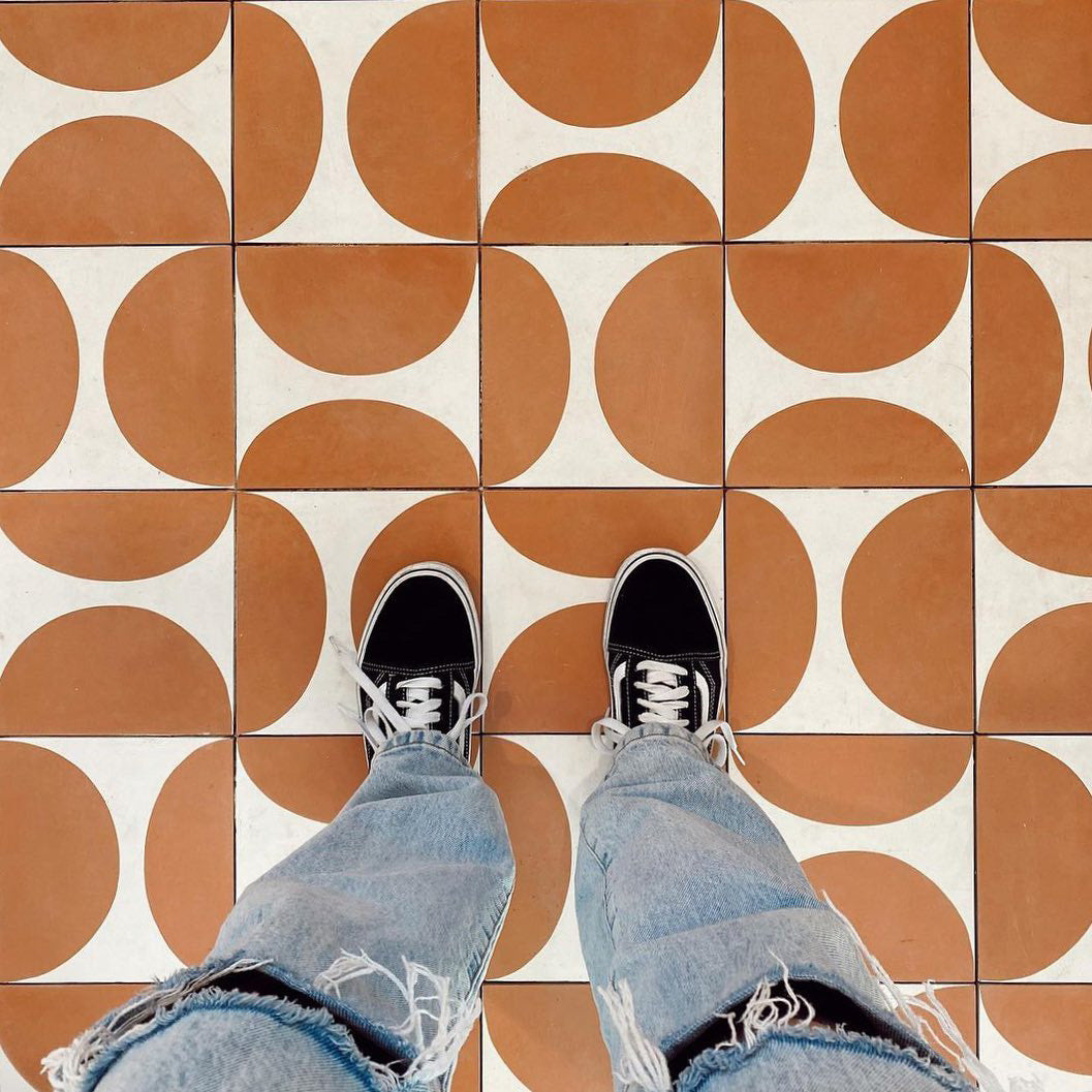 A person in ripped blue jeans and black sneakers stands on a tiled floor featuring Zia Tile's Pomelo Terracotta 4x4, Sample, with a bold brown and white geometric pattern. Photo taken from above, showing just their legs and shoes.