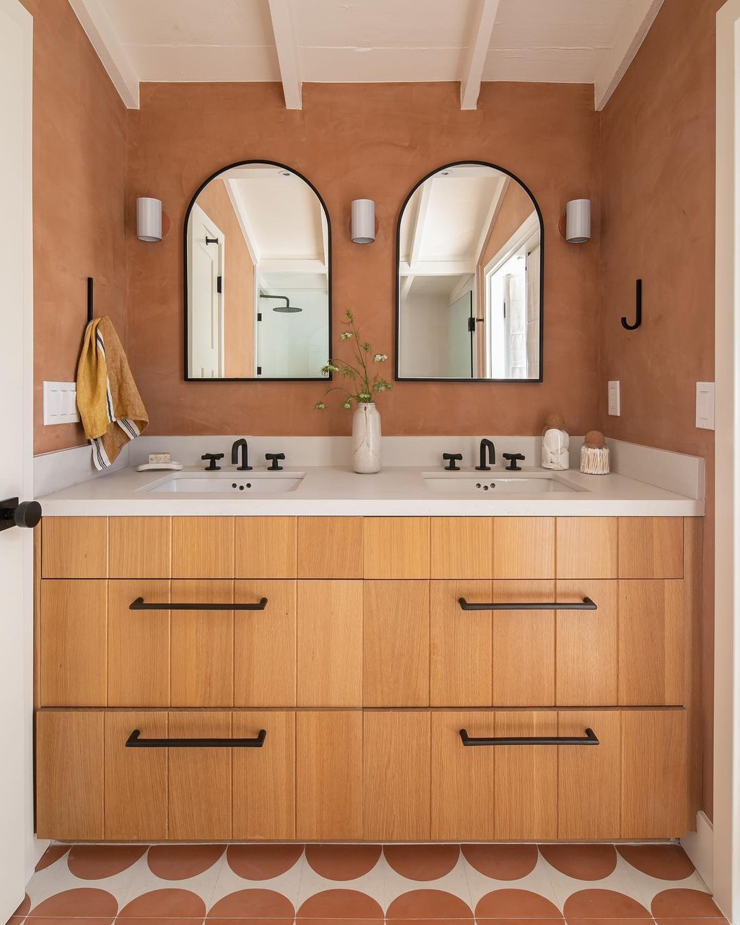 A modern bathroom features a wooden double-sink vanity, two arched mirrors, black fixtures, and terra-cotta Pomelo Terracotta 4x4 tiles by Zia Tile. A flower vase sits between the sinks, with towels hanging on the wall.