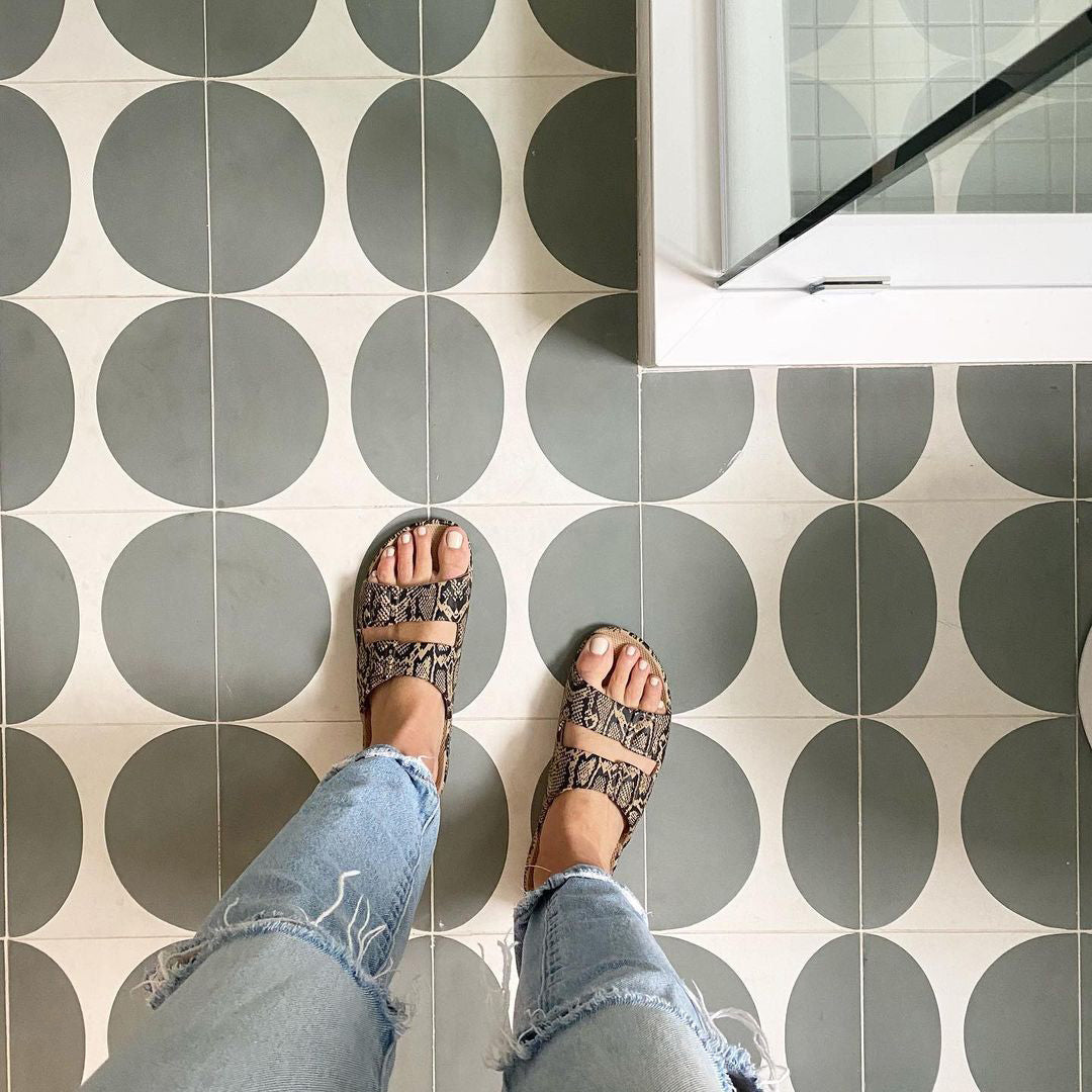 A person in ripped jeans and snakeskin sandals stands on a tiled floor featuring Zia Tile’s Pomelo Zeppelin 4x4 cement tile sample near the corner of a glass shower.