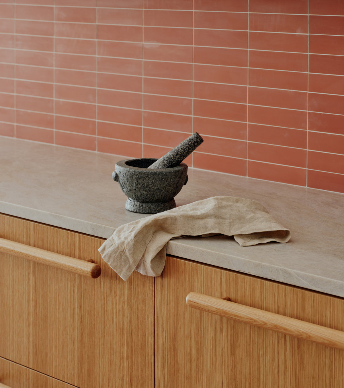 A stone mortar and pestle and a linen cloth on a kitchen counter in front of a terracotta-colored Zia Tile Pompeii 2x8 subway tile backsplash.