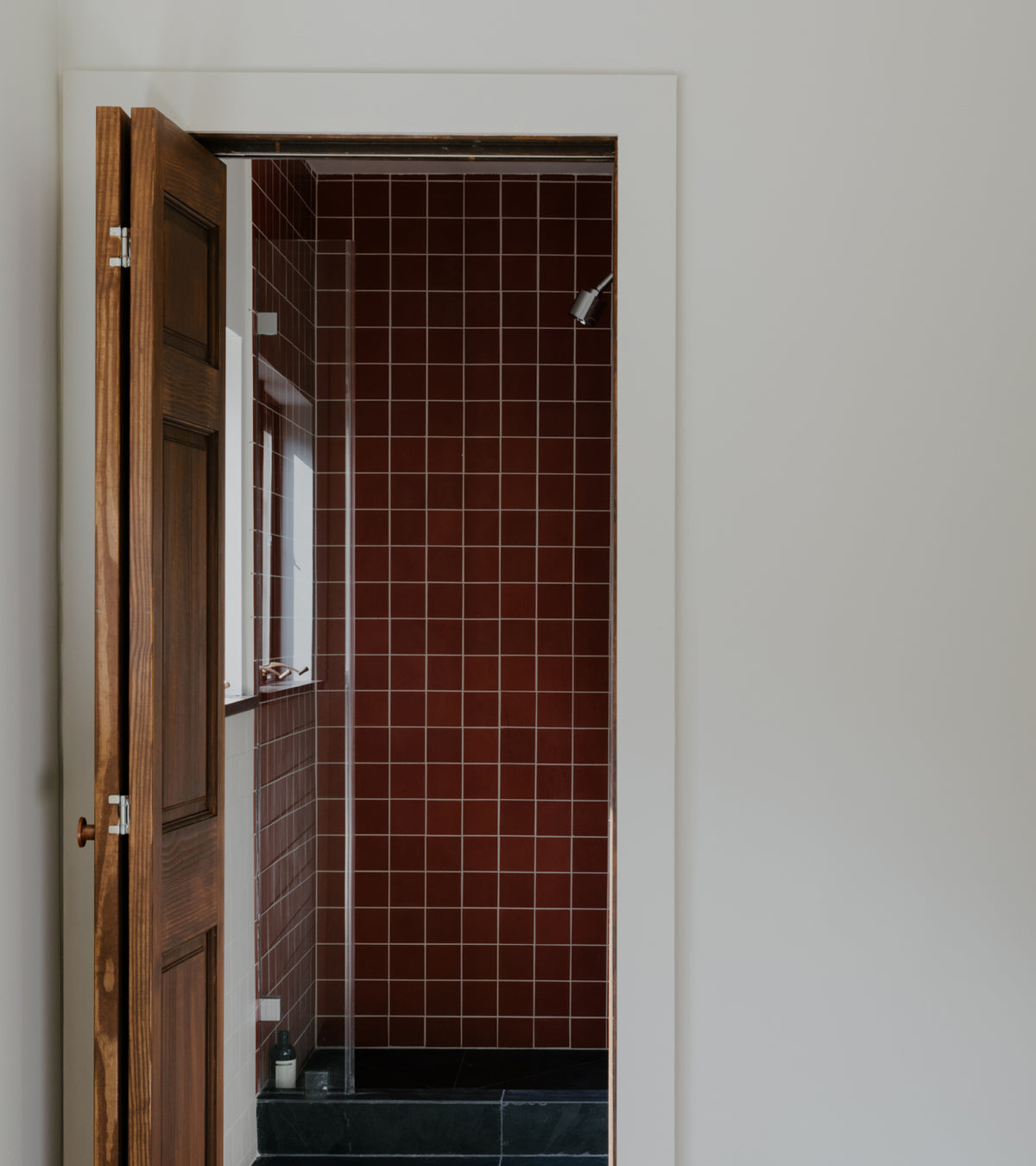 A view through an open wooden door into a shower stall featuring deep red Pompeii 4x4 cement square wall tiles by Zia Tile.