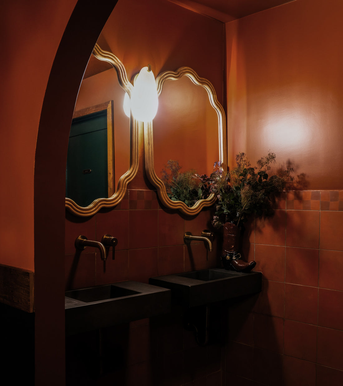 A dimly lit bathroom with orange walls, twin black sinks, gold-framed mirrors, brass faucets, and a boot-shaped vase sits atop Pompeii 8x8 handcrafted cement tile by Zia Tile under a single light.