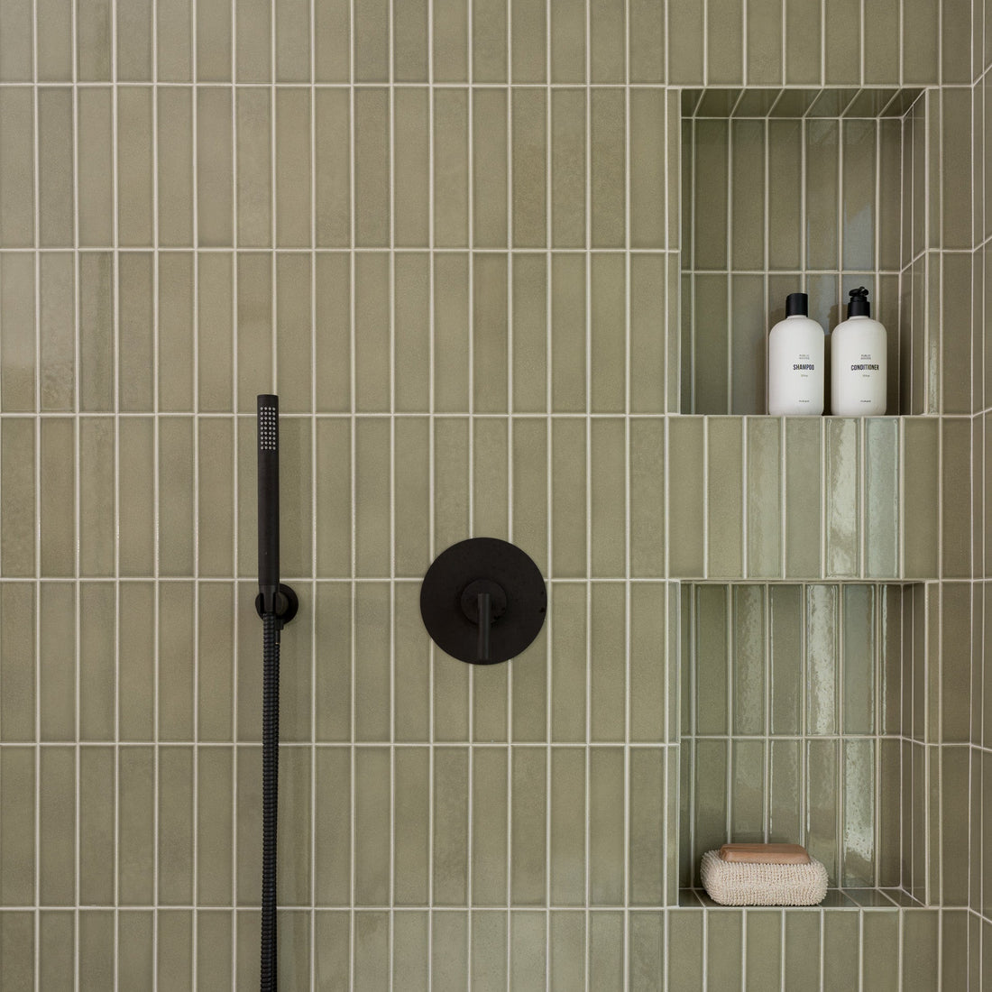 Shower wall clad in Zia Tile’s Ponderosa 2x8 olive-green vertical ceramic tiles, with built-in shelves for two bottles and a soap bar, plus matte black fixtures featuring a handheld shower and control knob.