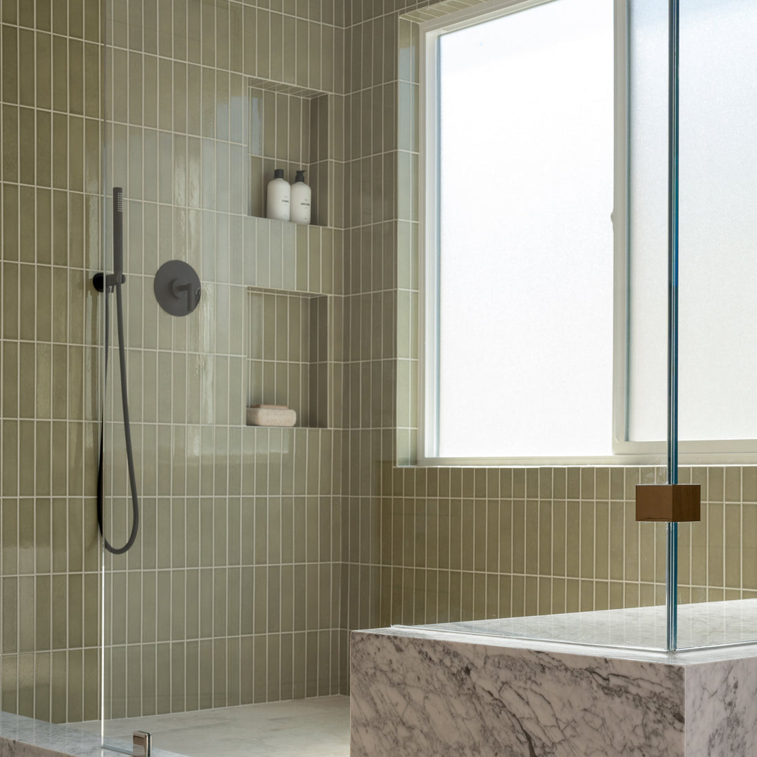 A modern shower features Zia Tile’s Ponderosa 2x8 ceramic tiles in light olive-green, matte black showerhead, built-in shelves, a large frosted window, and a marble bench in the foreground.