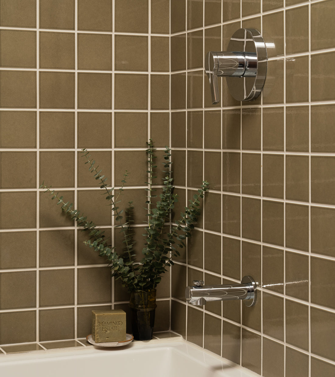 A modern bathtub corner features Zia Tile’s Ponderosa 4x4 glossy ceramic tile, a chrome shower handle, a small vase with eucalyptus, and a bar of soap on a dish resting on the tub edge.