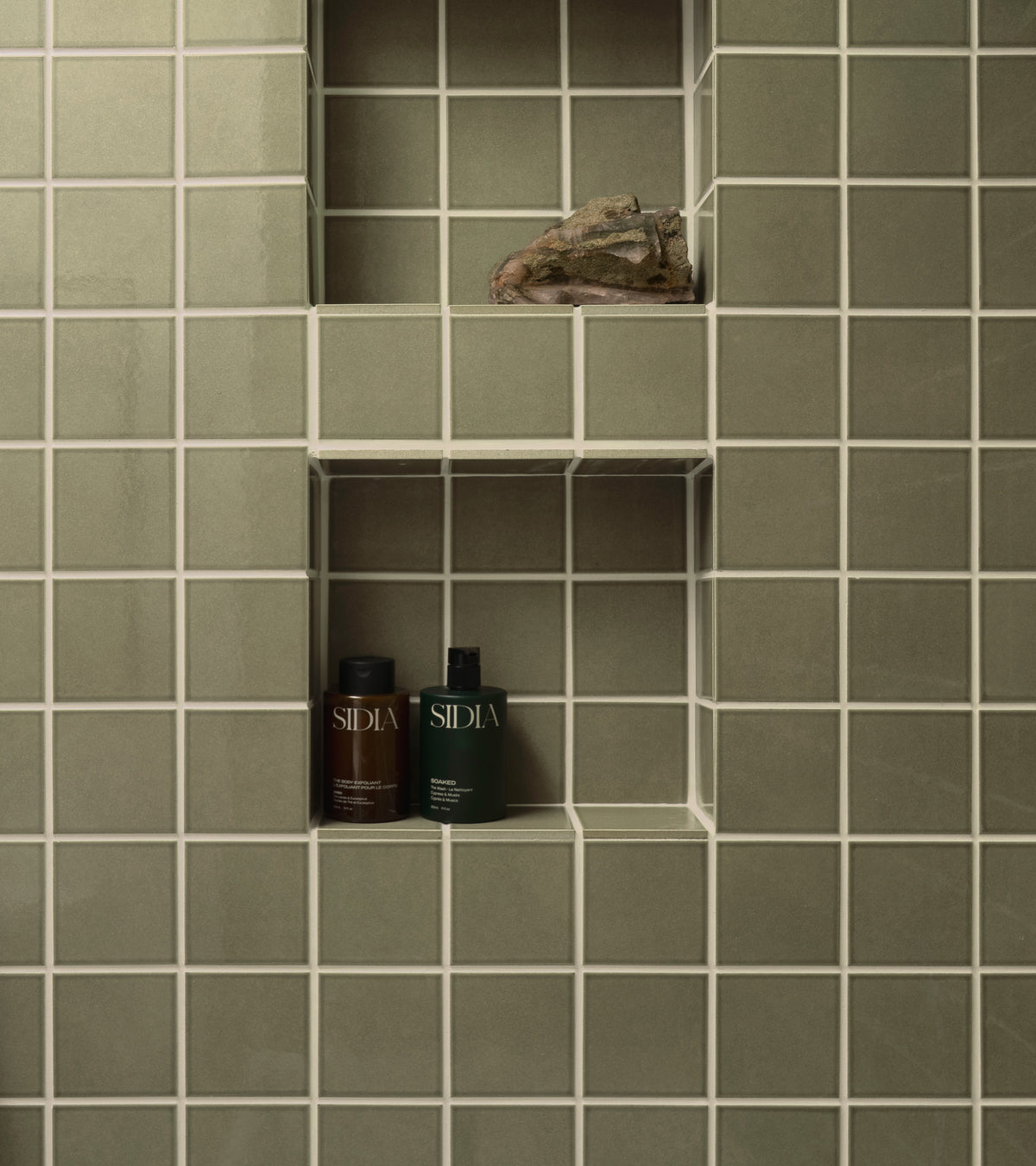 Sage green glossy Ponderosa 4x4 ceramic tiles with white grout on a bathroom wall featuring two recessed niches, one containing a rock and the other holding two bottles of skincare product.